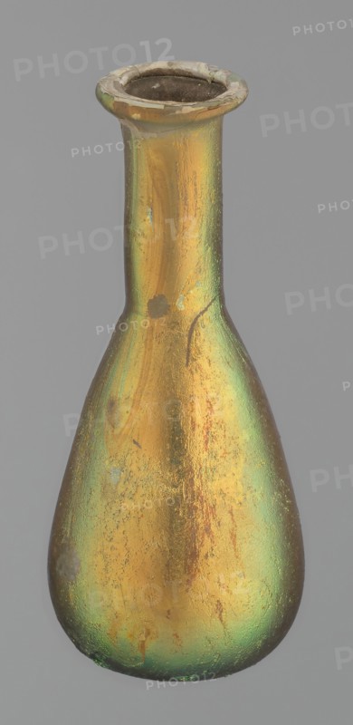 Flask, unknown eastern workshop, Goluchów Greater Poland Voivodeship, glass, antiquity, Roman Empire. Flask with a slender, pear-shaped body and a lower neck ending in a ring-shaped rim. Flat bottom. Covered with multicolored patina. 1st–2nd century, unknown type of site/place of origin, vessel, height 9.4 cm, A single elongated bottle with a narrow neck and flared rim sits against a plain gray background, its surface showing an iridescent sheen in gold and green tones with areas of darker brown and lighter worn spots, a few small rough patches and streaks visible on the body, and the interior of the rim appearing dark.