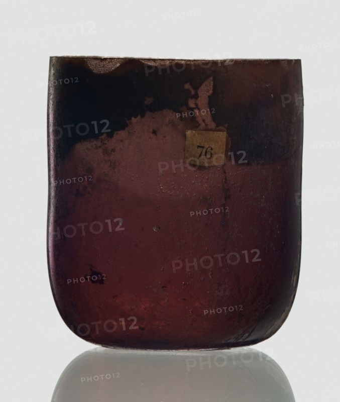 Bottle, unknown eastern workshop, Goluchów Greater Poland Voivodeship, 3rd century, vessel, height 4 cm, width 3.4 cm, A close-up of a rounded rectangular glass vessel with a smooth curved base, showing a translucent deep reddish-brown color that becomes darker near the top and edges, with areas of darker brown and almost black mottling and some lighter brown patches; the surface has small bubbles, pitting, and irregular texture, and there is a small aged paper-like square adhered near the upper center in a faded beige tone; the vessel sits on a reflective pale surface that shows a soft mirrored reflection, and the background is a very light, neutral color.