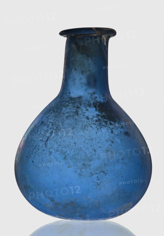 Bottle, unknown eastern workshop, 1st–2nd century, vessel, height 7.5 cm, width 5.5 cm, A rounded glass vessel with a narrow neck and flared rim, predominantly blue with areas of darker blue and blackish mottling and some lighter blue patches, showing a matte, weathered surface texture with small blemishes and spots; the rim has a darker edge with a small lighter chip, and the object sits on a reflective pale surface against a light gray background.