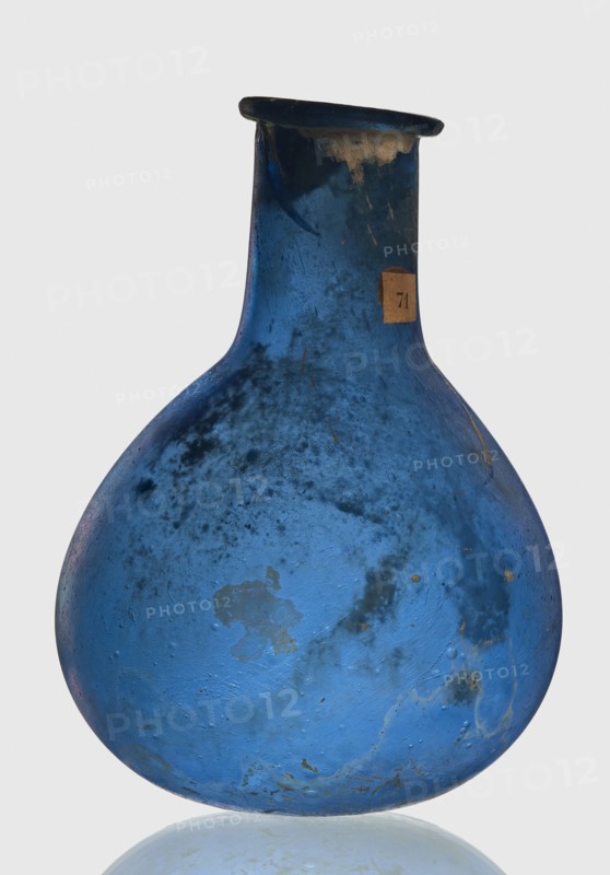 Bottle, unknown eastern workshop, Goluchów Greater Poland Voivodeship, 1st–2nd century, vessel, height 7.5 cm, width 5.5 cm, A single rounded glass vessel with a narrow neck and flared lip, shown against a pale background. The vessel is predominantly blue with areas of darker blue and some lighter blue patches. There are several mottled and cloudy dark spots and stains across the surface, along with a few small areas where the color appears worn or discolored to a tan or beige. The glass surface shows texture with tiny pits and irregularities and a few faint scratches. The overall shape is bulbous at the base tapering to a slim neck with a slightly uneven rim. The bottom edge sits on a reflective surface that produces a pale blue reflection.