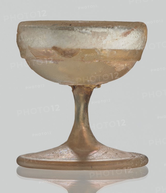 Cup, Egyptian workshop, Goluchów Greater Poland Voivodeship, 3rd–4th century AD, Roman Empire 27 BC–476, vessel, height 3.1 cm, width 3.2 cm, A small translucent vessel with a rounded bowl and a flared base sits against a pale gray background; the glass shows a mix of pale amber, light brown, and hints of green with areas of opalescent sheen and cloudy white patches, surface irregularities, and roughened edges; the stem is slender and slightly tapered connecting the bowl to a broad flat foot that has a reflective underside and subtle iridescent speckles in pink and purple tones.