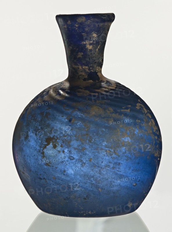 Bottle, unknown eastern workshop, Goluchów Greater Poland Voivodeship, 3rd–4th century, vessel, height 10 cm, width 3.8 cm, A rounded glass vessel with a short neck and flared rim, predominantly deep blue with irregular patches and speckles of darker tones and areas of brownish weathering, showing a mottled, uneven surface texture and subtle variations of lighter and darker blue across its bulbous body, photographed against a plain light background.