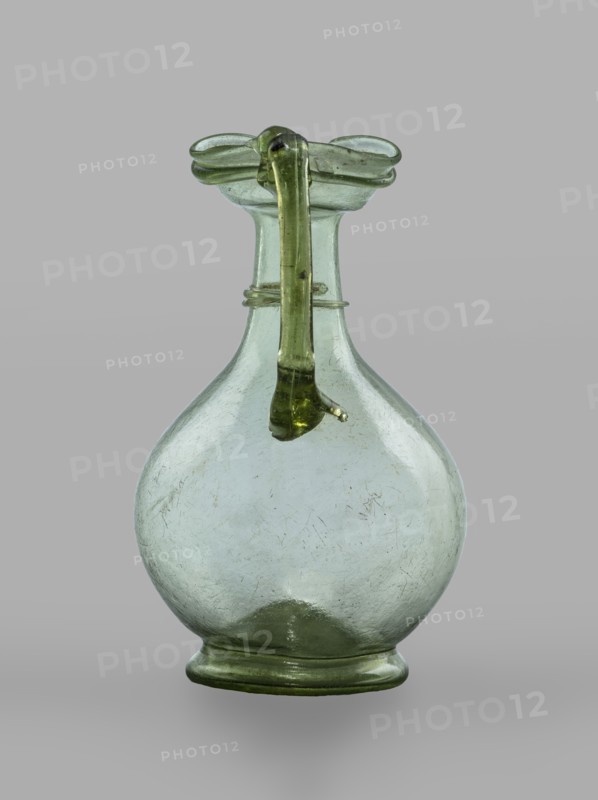 Oinochoe, Transparent, light green glass. Spherical belly on a foot with a clearance, created by pressing inward the central part of the bottom of the vessel; decoration on the cylindrical neck in the form of a single glass thread of the same color, wound horizontally. The handle is glued to the arms and extends in an arc to the edge of the trefoil rim; free-blown. 4th century, Cyprus; vessel oinochoe. Base diameter 4.8 cm, weight 0.062 kg. Type: Isings form 124a., A blown glass vessel with a rounded body and a narrow neck topped by a flared rim, featuring a looped handle that extends from the rim down toward the body; the glass appears translucent with green and pale green tones, and the surface shows visible scratches and small irregularities, while the background is a uniform light gray.