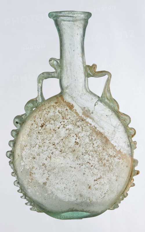 Ampoule, Egyptian workshop, 3rd century, Roman Empire 27 BC–476, vessel; height 12 cm, width 7.3 cm, A translucent glass vessel with a rounded, flattened body and a tall narrow neck, showing a pale green and clear glass color with areas of beige and brown surface encrustation; the body has a scalloped rim detail around its edges and two decorative looped handles near the neck that extend down onto the shoulder; the glass surface shows cloudy, pitted texture and a few visible hairline cracks.