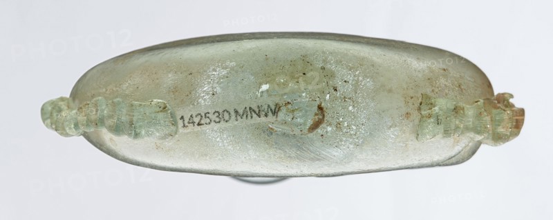 Ampoule, Egyptian workshop, 3rd century, Roman Empire 27 BC–476, glass vessel, height 12 cm, width 7.3 cm, An elongated, flattened glass vessel seen from the side with a translucent, pale green surface that shows areas of cloudiness and iridescent sheen, along with patches of brownish discoloration and small dark speckles; a pair of ornate, twisted handles of the same pale green glass project from opposite ends, showing similar weathered texture; the background is a uniform light gray.