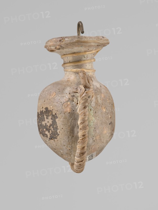 Ampoule, Egyptian workshop, 4th century BC, vessel, height 5.2 cm, width 4.2 cm, An aged, bulbous ceramic vessel with a rounded body and a flared neck, shown against a plain light gray background; the vessel surface is mottled with shades of beige, tan, brown, and hints of pale green, and shows areas of darker brown patina and spots of weathering; a thick twisted handle runs from near the neck down the side to the base, the handle appearing textured and beige with darker accents; the rim is irregular and slightly worn, with concentric ridges at the neck that are highlighted in a pale tan; a small metal hook protrudes from the top of the image, showing a dark brown, slightly corroded surface.