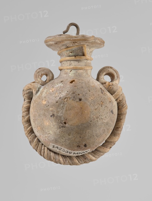 Ampoule, Egyptian workshop, 4th century BC, vessel, height 5.2 cm, width 4.2 cm, A rounded ceramic vessel displayed against a plain gray background, showing a bulbous body with a narrow neck and a flared rim; the surface of the vessel is mottled with beige, tan, light brown, and small darker brown speckles, with areas of lighter cream and pale gray; two looped handles rise from the shoulder, each with a coiled rope-like attachment that curves down and around the base, the rope-like element appearing in similar beige and tan tones with shadowed ridges; the rim has a hooked metal loop inserted, which is dark brown with a slightly rusty patina; the vessel surface shows small pitted holes and textured wear in various spots, and a slightly darker circular area is visible on the front of the body.