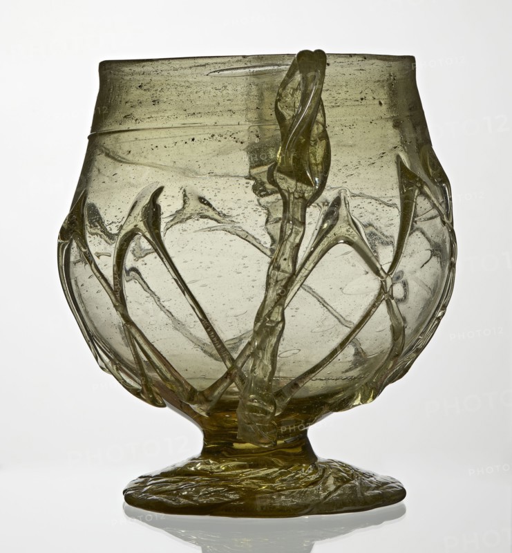 Skyphos, deep bowl with wide foot and two small vertical handles. Surface decorated with a zigzag ornament applied with irregular threads. 3rd–4th century AD, Roman Empire 27 BC–476. Vessel, height 11 cm., A translucent glass vessel with a rounded bowl and a short stem on a flattened foot, primarily pale olive-green in color with darker green accents; the surface shows irregular bubbles and streaks, and several raised twisted glass ribbons and arc-shaped applied elements wrap around the bowl creating a decorative network; the rim is slightly uneven and the base appears textured with rippled glass, all shown against a light neutral background.