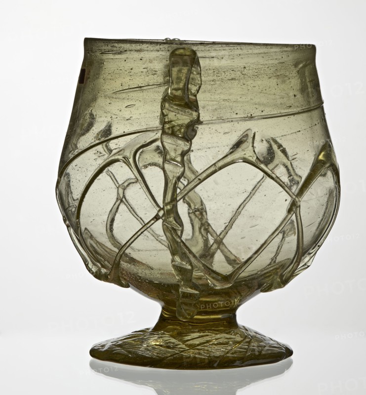 Skyphos, Deep bowl with wide foot and two small vertical handles. Surface decorated with a zigzag ornament applied with irregular threads. 3rd–4th century AD, Roman Empire 27 BC–476, vessel, height 11 cm, A translucent glass vessel with a rounded bowl and a flared rim sits on a flattened foot; the glass is a pale olive-green color with visible bubbles and streaks, and raised twisted trails of the same glass wrap around the body forming an irregular lattice. The surface shows a few rough areas and small inclusions, and the foot has a textured pattern in the same green tone. The background is a plain light color that contrasts with the olive-green glass.