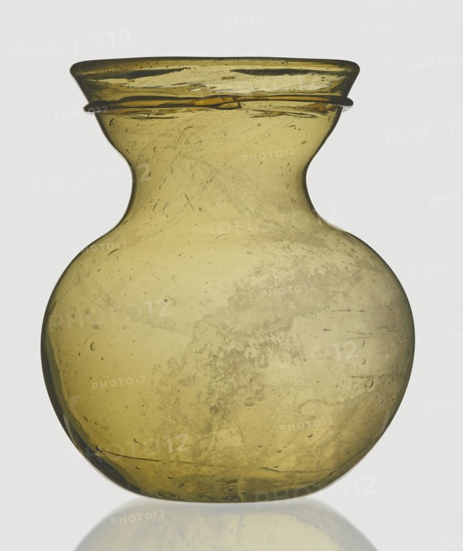 A viceless vessel, Goluchów Greater Poland Voivodeship, below the edge a glass thread is applied horizontally. Unspecified place of origin, vessel, height 12.4 cm, width 10.7 cm, A translucent rounded glass vessel with a narrow neck and flared rim, shown against a light background; the glass is an olive-greenish yellow with variations in tone, areas of cloudiness and internal bubbles, and a few darker green streaks and speckles visible throughout the surface, with a subtle reflection beneath it.