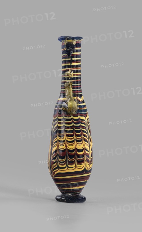 Amphoriskos; with an elongated neck; eastern Syrian or Cypriot workshop;, 2nd–1st century BC. Type: Grose group II:E, form III:4. Opaque glass with dark blue-green body and decoration of festoon patterns made with yellow and brown threads; discoidal rim; neck widening slightly downwards; baggy belly; solid foot. Vertical vices one partially chipped and double vices made of translucent light honey-green thread; formed on the core. Outer rim diameter 2.25 cm; height 14.80 cm; body diameter 4.10 cm; bottom diameter 2.85 cm. Vessel., A tall narrow vase with a rounded base and a small flat foot stands against a plain gray background; the vase is decorated with a repeating wavy stripe pattern in black, yellow, red, and a few dark blue bands, and it has a small applied loop handle near the neck in a translucent yellowish color, with the neck flaring slightly at the rim which shows dark blue and yellow tones.