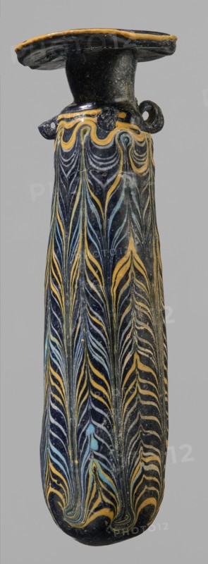 Alabastron, cylindrical belly, 4th–3rd century BC, vessel alabastron, height 17.4 cm, width 4.4 cm, origin unknown., A tall narrow vessel photographed against a neutral gray background, with a flattened rim and short neck leading to a long tapering body; the surface is predominantly dark, glossy black with decorative flowing featherlike patterns in yellow and light blue that run vertically along the body, and a band of undulating yellow lines near the shoulder; the rim and small looped handles show black with yellow edging, and the overall appearance is of a glazed, slightly worn object with fine linear motifs and subtle surface speckling.