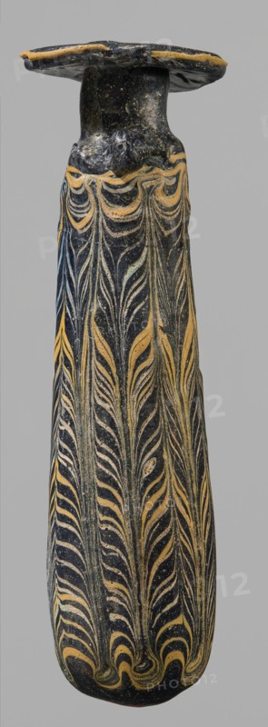 Alabastron, cylindrical belly, 4th–3rd century BC, unknown origin, vessel alabastron, height 17.4 cm, width 4.4 cm, A tall narrow object with a flared flat top, predominantly dark in color with decorative flowing featherlike patterns in a yellowish tone running vertically along its length, showing areas of lighter and darker speckling on the dark surface and some subtle bluish highlights, the top edge also showing a rim of the yellowish color against the dark background.