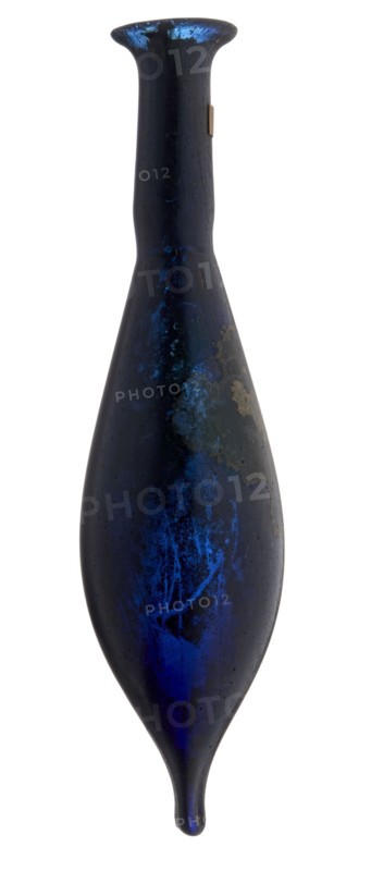 Vial, unknown eastern workshop, elongated belly with a pointed bottom, cylindrical neck and flared spout. Navy blue glass. 4th century, vessel, height 13 cm, width 1.8 cm, A tall, narrow vase-like object with an elongated teardrop shape and a flared rim, shown against a white background; its surface is predominantly dark blue to nearly black with areas of vivid metallic blue highlights and patches of lighter blue; there are a few irregular pale grayish or tan worn spots and mottling on one side; the form tapers to a rounded point at the bottom and has a smooth, slightly reflective texture overall.