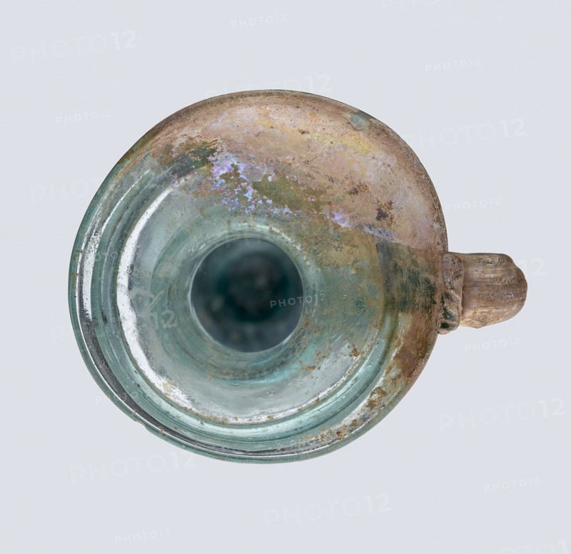 Bottle; eastern workshop, 3rd–4th century AD. Transparent bluish-green glass, free-blown; short cylindrical neck and funnel-shaped rim with two applied glass threads in a ring beneath the rim. Outer diameter of rim: 5.20 cm. Height: 15 cm. Belly diameter: 5.30 cm. Bottom diameter: 1.65 cm. Vessel; height 15 cm; width 5.3 cm; weight 0.066 kg., A circular glass vessel viewed from above with a rounded rim and a short handle on one side, showing a mix of greenish-blue and pale turquoise glass tones, with areas of brownish tan and iridescent purplish sheen across the upper surface, patches of dull green and muted yellowish discoloration, and some cloudy and weathered textures on the interior and rim.