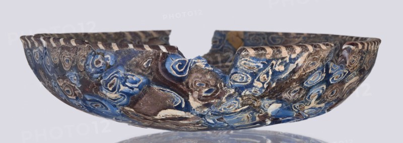 Bowl, Egyptian workshop; mosaic glass, glass vessel with floral patterns. Bowl with a flat bottom whose walls widen evenly toward the top then narrow at the rim; cut flat at the top. Made of thick glass using the mosaic technique; floral patterns in blue, purple, yellow, green, and white. Type: Isings form 1. Semicircular bowl; sagging on the form; 1st century, Roman Empire 27 BC–476. Vessel; height 3 cm, width 13.5 cm., A shallow, broken bowl with an irregular rim showing a jagged gap, decorated with swirling, concentric patterns in shades of blue, brown, beige, and white; the surface shows areas of glossy and matte texture with worn and rough patches, and the interior and exterior share the same patterned design, all set against a pale gray background with a subtle reflected shadow beneath.