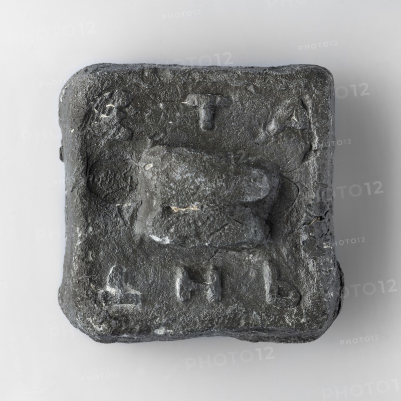 Square weight with diagonally cut side edges. The upper surface is decorated with a plastic representation of an astragalus. At the upper edge are horizontally arranged convex letters ETA, at the lower edge THb. A second stamp with an image of a sea turtle was impressed next to the astragalus. 6th century BC, Attica. Weight 2 trade mines, unknown Attic workshop. Weight: 1205 g; height 7.1 cm; length 6.9 cm; width 2.6 cm., A roughly square, dark grey object with an uneven, textured surface sits against a light grey background; the object has rounded corners and a raised, irregular central shape that projects from the flatish face, and there are a few shallow depressions and raised marks around the central form, with small patches of lighter grey scattered across the surface.