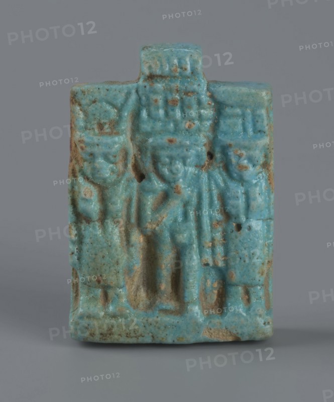 Amulet – plaque with a triad of deities: Harpocrates Horus, Isis, Nephthys. Amulet in the shape of a plaque with a triad of deities standing on a base: central figurine of Horus, Isis at left, Nephthys at right. Back of plaque flat; pendant above the upper edge. Date: 664–30 BC, Late Period–Ptolemaic Period probable. Country of find: Egypt. Material/function: amulet. Dimensions: height 3.3 cm, width 2.2 cm, depth 0.7 cm., A small rectangular turquoise-blue carved object with a slightly rough, speckled surface and patches of brownish discoloration, showing a raised central figure flanked by two smaller raised figures; the top has a short rectangular projection, and the lower edge is slightly worn with visible areas of exposed material; the background and base are a neutral gray.