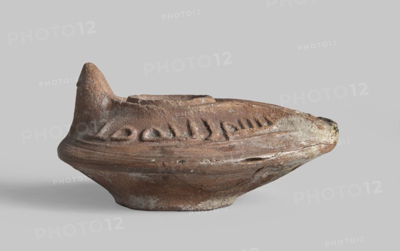Oil lamp, Oval lamp from a mold. Gently sloping arms with a Kufic inscription bismi l'lahi mina l'lahi = in the name of Allah, from Allah. A large oil opening surrounded by a convex shaft. A second outer shaft also surrounds the burner channel, on which another inscription one word is inscribed. Conical nozzle. Ring-shaped, round base base-ring, in the center of which there are Arabic letters l, r, k?. Islamic art; Arabic ceramics; utility ceramics; Arabic inscriptions; Arabic language; everyday life in the Middle East. 8th–9th century, Syro-Palestine. Materials: ceramics. Function: oil lamp. Dimensions: height 4.4 cm, length 9.1 cm, width 6.4 cm., A weathered ceramic object rests on a neutral gray surface and background, showing a rounded base that flares outward into a wider midsection and tapers to a pointed protrusion at one end and a raised spout-like rim at the top. The surface texture is rough and pitted with visible chips and cracks, and there are shallow carved or impressed gr...