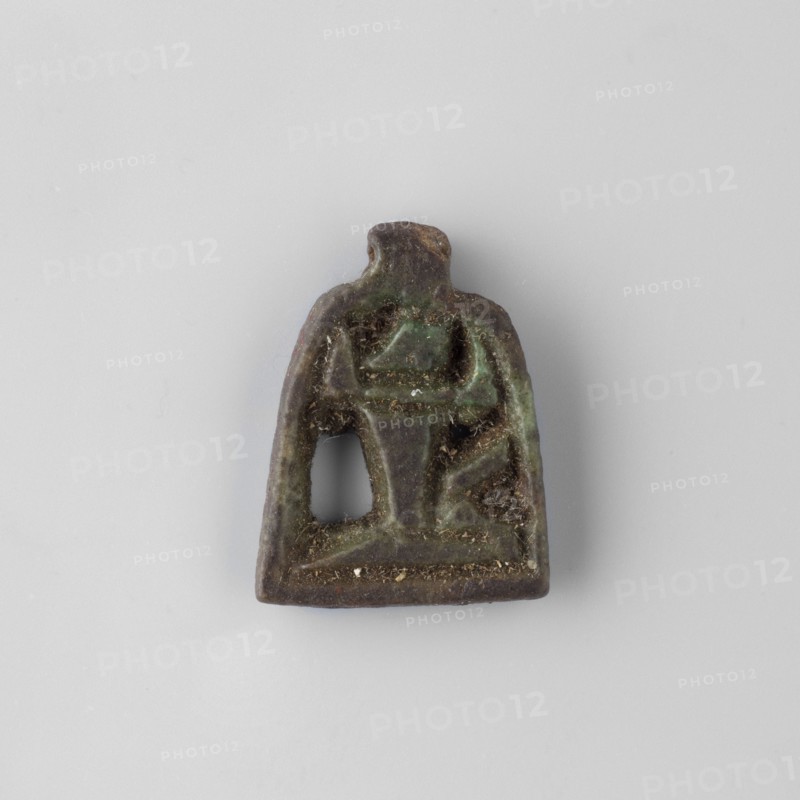 Amulet – plaque with the god Heh; unknown creator; Shu mythology. Openwork plaque showing the god Heh depicted in a very schematic manner. The palm stems held by the figure form a kind of frame with a pendant. 664–332 BC Late Period, Egypt. Amulet; height 1.9 cm, width 1.4 cm, depth 1.8 cm., A small worn object with an irregular trapezoidal outline and a rounded top, showing a raised central motif and a hollowed opening on one side, sitting on a plain light gray background; the object displays mottled dark brown and green tones with areas of lighter green patina and small specks of beige debris.