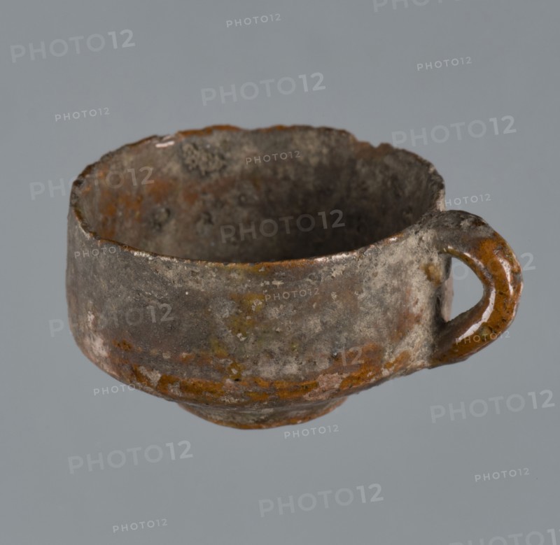 Miniature scoop with a flat bottom; walls widening diagonally to 1/3 then running vertically upwards; single vertical cylindrical handle. Unspecified maker; origin: Greece. Vessel/scoop. Height 1.5 cm, width 2.5 cm., A small cup with a rounded body and a loop handle, showing a rough, pitted surface and uneven rim; the cup displays a mottled palette of brown, gray, orange, and a few greenish areas, with darker interior tones and subtle patches of lighter off-white on the exterior.