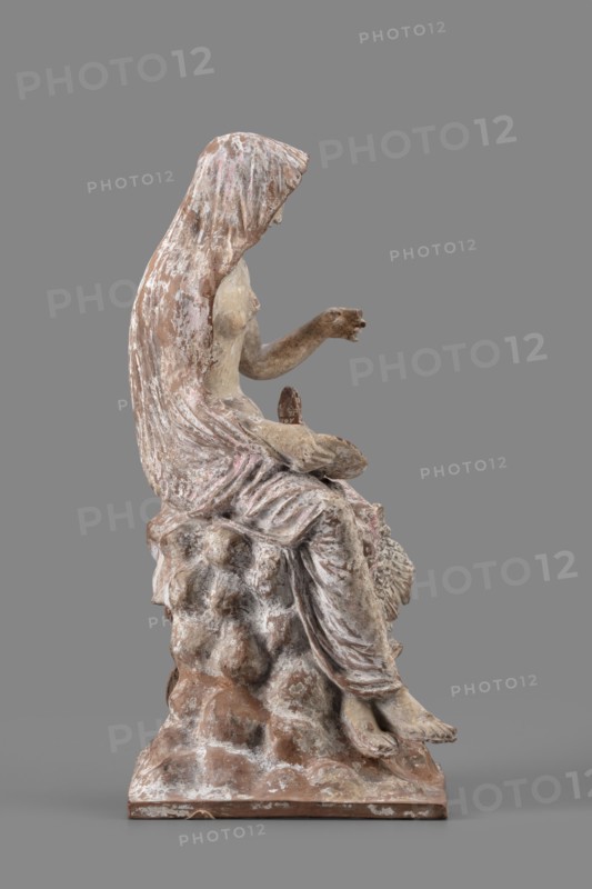 Figurine of Leda with a Swan, A woman sits on a rock and feeds a swan standing next to her. She is dressed in a cloak thrown over her head and back, which falls along her body and completely envelops her legs, leaving the rest of her body exposed. In her right hand, resting on her knees, she holds a platter. With her left hand, bent at the elbow and extended, she holds it above the swan, which rests on her exposed hip, its head extended towards her. The woman bows her head and looks at it. The swan has outstretched wings. The woman's sandaled feet do not rest on the rock but dangle. The square base forms one with the rock. Cloak painted pink. Mid-3rd century BC, Myrina; figurine; height 24 cm, width 15 cm, depth 12 cm, weight 0.56 kg., A weathered sculptural figure seated on a rough, rock-like base, shown in profile facing right, with a draped hood or cloak falling over the head and back; the figure's exposed arm is extended forward with the hand slightly open, and the legs are relaxed...