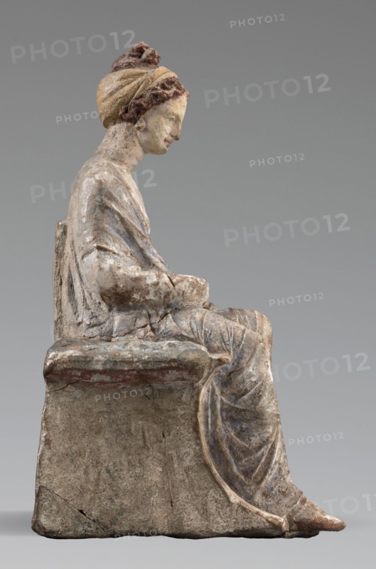Figurine of a woman. A girl sitting in a chair is dressed in a soft white chiton over which a blue, in some places lilac, cloak is thrown, which opens at the front. She rests her left hand on the chair cushion, while her right rests on her knees and is covered by the cloak. Her brown hair is parted at the front, combed upwards and girded. The full chair with forward-curved legs has a narrow and low backrest; the seat cushion is blue on top and red underneath. No base. Early 3rd century BC, Tanagra Greece. Terracotta figurine, height 16 cm, width 8 cm, depth 10 cm, weight 0.25 kg., A seated sculpture of a robed figure shown in profile, with a headwrap and hair detail, sitting on a block-like seat; the figure's robe has visible folds and drapery, and the hands rest in the lap. The surface shows weathering and wear with areas of rough texture and small cracks. Visible colors include muted beige and off-white on the skin and seat, pale yellow and brown tones in the headwrap and hair, faded...