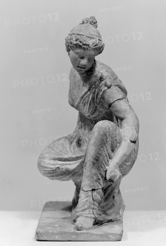 Terracotta figurine of a girl throwing dice. Figurine of a crouching girl throwing dice: the weight of the body rests on the left leg, bent at a right angle. She wears a long, finely pleated, sleeveless chiton. Hair tied in a bun with a wide headband kredemnos. Earring visible in left ear. Entire figurine rests on a rectangular base. Preservation: missing the right arm; glued in several places head, left arm from 2 fragments, robe, base from 3 fragments; part of the base filled with clay and plaster. Date: 4th–3rd century BC. Origin: Tanagra Greece. Material: terracotta. Type: figurine. Dimensions: height 13 cm, width 9 cm, depth 4.5 cm. Maker: unknown., A grayscale photograph of a sculpted figure of a seated woman on a rectangular base, shown from a slightly elevated frontal angle; the figure wears a draped garment with visible folds and appears to be adjusting or holding the drapery near a bent knee, with one arm extended downward and the other arm resting near the torso; the head is...
