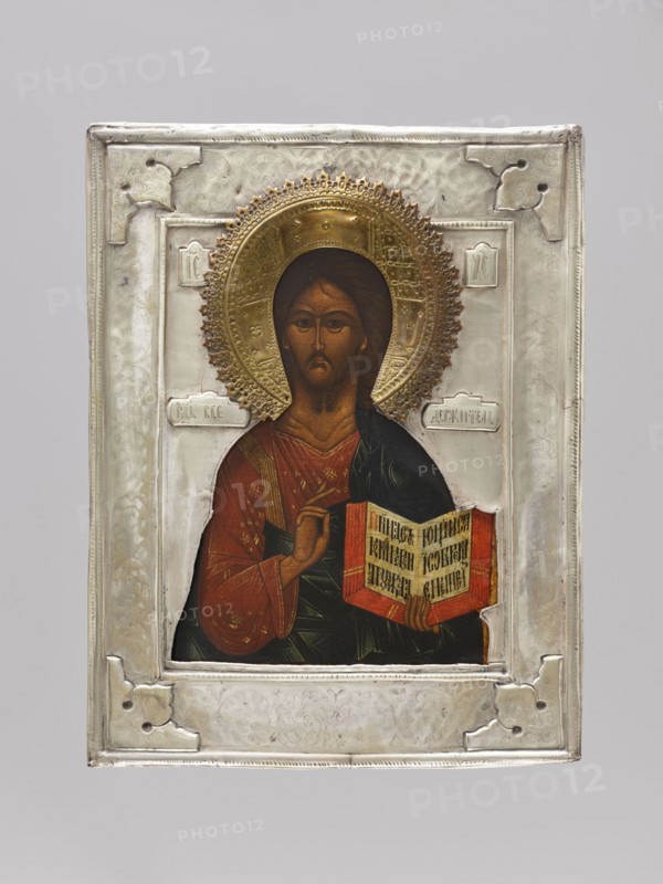 Icon in a cover: Christ Pantocrator. The icon represents a traditional, half-length variant of Christ Pantocrator referring to designs dating to the 16th century. The Savior's face is elongated, ascetic, with a light brown complexion brightened by tones; lower eyelids, forehead wrinkles, and neck folds are clearly marked. Christ wears a red chiton decorated with fine gold ornaments, an ochre sash covered with assyst, and a navy blue himation with rather small, pointed folds. The inscription on the book is made with a decorative ribbon. Around Christ's head is a cross halo with the letters OOH. On both sides of the figure are inscriptions: the Christological monogram and Lord Almighty. The entire field, the icon background, and the halo are covered with a brass cover with an applied wreath. 18th century, Russia; icon with cover; height 31 cm, width 24 cm., An icon painting of a figure with a golden halo set within a silver frame, showing a person with brown hair and a brown beard wearin...