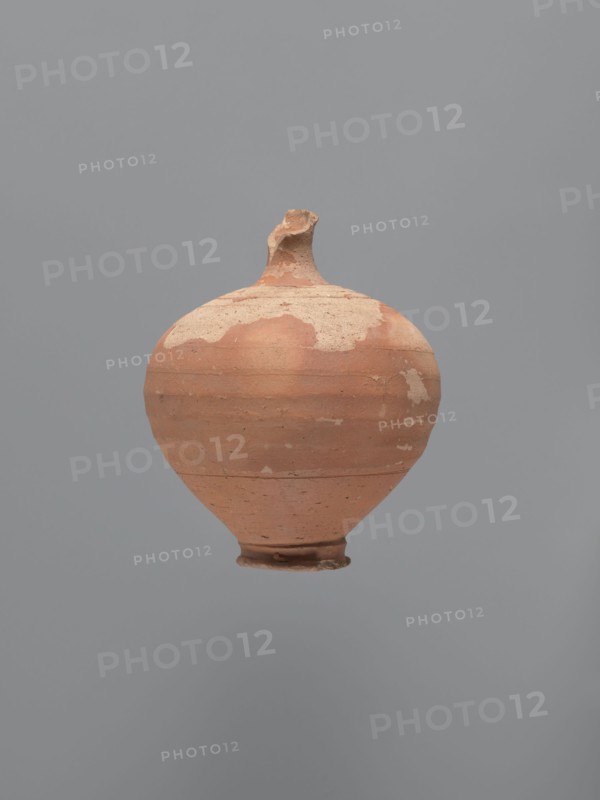 Jug, unknown Aswan workshop. Jug with a rounded rim, a narrow and upwardly flaring neck with a ring-shaped flange. Spherical body with a large ribbon-shaped handle glued to the lower part of the body and the middle part of the neck. Ring-shaped foot. Traces of turning slightly smoothed. Exterior covered with red-brown engobe, interior beige-pink, even. Kaolin ceramic mass Gempeler type IA. Egyptian sigillata, Egyptian pottery, Coptic vessels, tableware. 5th–6th century. Edfu find location. Vessel, jug; height 14.1 cm; lip diameter 2.4 cm; neck diameter 1.8 cm; body diameter 11.4 cm; base diameter 4.6 cm., A rounded clay vessel with a narrow base and a short, irregular neck suspended against a plain background; the vessel shows worn and flaked surfaces with patches of lighter and darker terracotta tones, a few areas of pale beige residue, and a generally matte, textured appearance in shades of reddish-brown, beige, and muted orange against a solid gray backdrop.