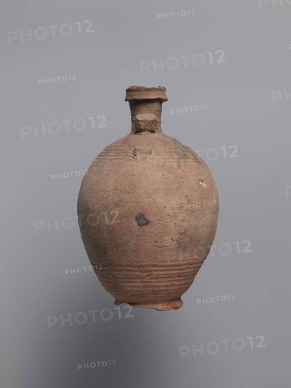 Bottle, unknown Egyptian workshop. Bottle with a rounded and falling rim, concavely profiled. Almost cylindrical neck flows into rounded shoulders covered with fine ribs. Two handles one fragmentary are attached to the lower part of the neck and upper part of the shoulders. Ovoid body with grooves on the lowest part traces of turning; ring-shaped foot. Outer surface brown and burnished. Made of A4 marl clay with limestone inclusions. Faint painted decoration on the shoulders in black paint: two arcades composed of parallel arches separated by a row of dots, arranged symmetrically one between the handles. Irregular concentration of a black substance at the highest bulge of the body below a damaged handle, probably bitumen used to seal or repair the wall. Painted Coptic pottery, painted Coptic vessels; marl clay; bitumen. 6th century, Edfu. Vessel bottle. Height 22.7 cm; spout diameter 4.3 cm; neck diameter 3.0 cm; belly diameter 14.9 cm; bottom diameter 7.2 cm., An aged rounded clay ves...