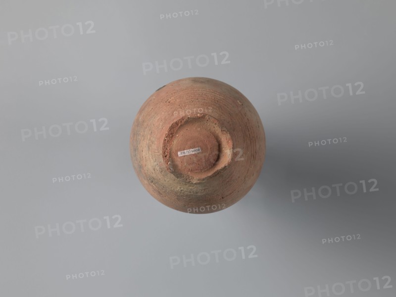 Bottle, unknown Egyptian workshop. Bottle with a rounded and falling rim, concavely profiled. The almost cylindrical neck flows into rounded shoulders covered with fine ribs. Two handles, one fragmentarily preserved, are glued to the lowest part of the neck and the lower part of the shoulders. The ovoid body is grooved on its lowest part traces of turning; the bottom is a ring-shaped foot. Outer surface brown and burnished. Made of A4 marl clay with limestone inclusions. On the shoulders is faint painted decoration in black: two arcades composed of parallel arches separated by a row of dots, arranged symmetrically, one between the handles. At the highest bulge of the body, below the damaged handle, is an irregular concentration of a black substance, probably bitumen used to seal or repair the wall. 6th century, Edfu. Vessel bottle. Height 22.7 cm; spout diameter 4.3 cm; neck diameter 3.0 cm; belly diameter 14.9 cm; bottom diameter 7.2 cm., A round, weathered clay pot viewed from above,...