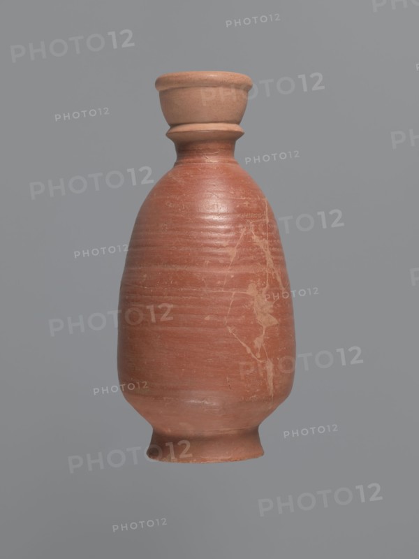 Bottle, unknown Aswan workshop; baggy-shaped bottle without preserved spout. The neck widens toward the top with a circumferential bulge and another at its base. Sloping shoulders; belly is baggy, flowing into the base on a low foot. Pink kaolin ceramic mass Gempeler IA. Partially smoothed turning marks visible on the surface. Outer walls covered with polished red engobe. Egyptian sigillata/Coptic pottery; Egyptian imports in Nubia. 2nd half 5th–1st half 6th century; find from Edfu. Vessel bottle. Height 20.6 cm; diameter 3.4 cm neck, 11.3 cm body, 7.7 cm bottom., A single pottery vessel centered against a solid gray background, with an elongated rounded body tapering to a narrow neck and flared rim. The vessel shows a smooth, slightly worn surface with visible horizontal turn lines and a few irregular pale streaks and blemishes on its body. The colors visible are terracotta, light brown, pale beige, and gray from the background. The rim and base appear slightly lighter in color than t...