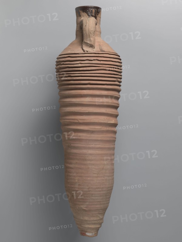 Amphora, unknown Egyptian workshop; amphorae, Egyptian amphorae, Egyptian ceramics, storage vessels, Coptic vessels, amphorae; Nile clay. Wine transport vessel with a spindle-cylindrical, long body. Steep arms, cylindrical neck ending with a straight rim. Cylindrical viscæ, oval in cross-section, are attached to the lower part of the arms and to the middle part of the neck. The entire body has grooves of varying density and size; the densest are in the lower and upper parts. No cylindrical foot. Brown, alluvial clay Naqlun AC1A ceramic type. Outer surface brown and even. Interior and the upper outer neck coated with a black resinous substance. 2nd half of the 4th–5th century. Findspot: Edfu. Vessel; amphora. Height 65 cm; rim diameter 7 cm; body diameter 19 cm., A tall, elongated pottery vessel with a narrow neck and a single handle attached near the top, featuring repetitive horizontal ridges along most of its body. The surface shows an earthy, muted terracotta color with variations o...