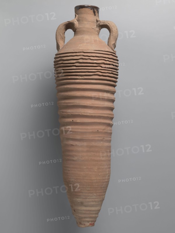 Amphora, unknown Egyptian workshop; amphorae, Egyptian amphorae, Egyptian ceramics, storage vessels, Coptic vessels, amphorae, amphorae, Nile clay. Wine transport vessel with a spindle-cylindrical, long body. Steep arms, cylindrical neck ending with a straight rim. Cylindrical vises, oval in cross-section, are attached to the lower part of the arms and to the middle part of the neck. The entire body has grooves of varying density and size; the densest are in the lower and upper parts. No cylindrical foot. Brown alluvial clay Naqlun AC1A ceramic type. Outer surface brown and even. The interior and the upper outer surface of the neck are coated with a black resinous substance. 2nd half of the 4th–5th century, Edfu find location. Vessel, amphora; height 65 cm, rim diameter 7 cm, body diameter 19 cm., A tall, narrow ceramic vessel with a rounded shoulder and a tapered base, featuring a pair of curved handles near the neck; the surface shows a series of horizontal ridges and grooves wrappin...