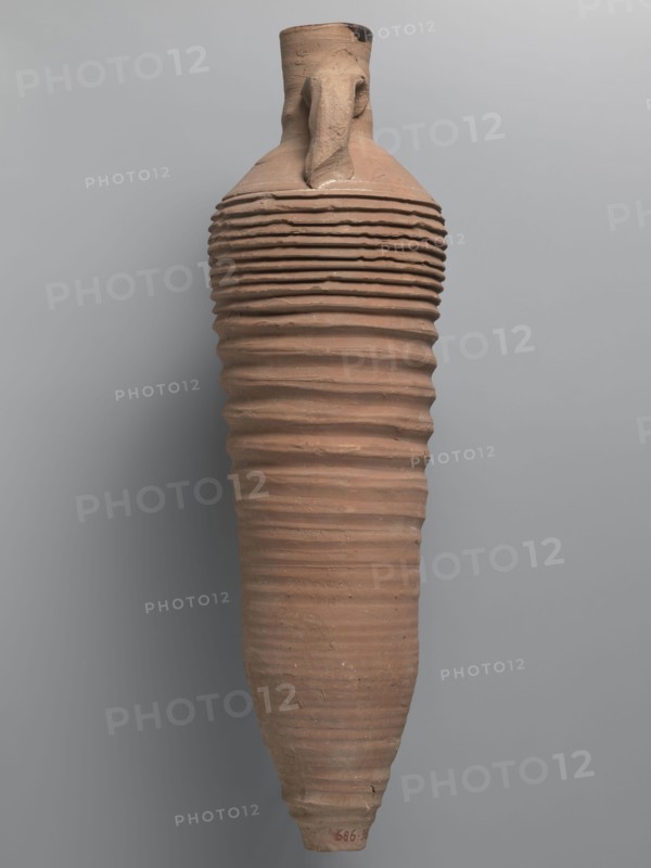 Amphora, unknown Egyptian workshop; amphorae, Egyptian amphorae, Egyptian ceramics, storage vessel, Coptic vessel, amphora; Nile clay. Wine transport vessel with a spindle-cylindrical, long body. Steep shoulders, cylindrical neck ending in a straight rim. Cylindrical vises, oval in cross-section, are attached to the lower part of the shoulders and to the middle part of the neck. Entire body has grooves of varying density and size; the densest are in the lower and upper parts. No cylindrical foot. Brown, alluvial clay Naqlun AC1A ceramic type. Outer surface brown and even. Interior and the upper outer surface of the neck coated with a black resinous substance. Dated 2nd half of the 4th–5th century. Findspot: Edfu. Vessel amphora. Height 65 cm; rim diameter 7 cm; body diameter 19 cm., A tall, elongated ceramic vessel with a narrow neck and a single handle shows a ribbed surface of uneven horizontal ridges extending along its body; the vessel is primarily terracotta brown with subtle vari...