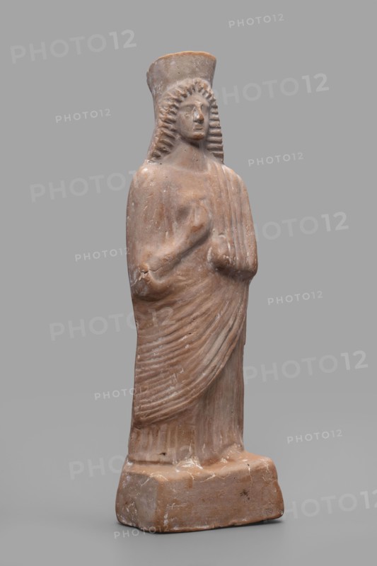Figurine of a woman with a polos on her head, unknown Boeotian workshop, female figurine, 475–450 BC, Boeotia, sculpture; height 26 cm, width 9 cm, depth 7 cm, weight 0.43 kg., The image shows a standing terracotta statue of a robed figure against a plain gray background. The figure has a tall, rounded headdress and stylized, ridged hair framing the face, with a calm, neutral expression. The body is wrapped in draped garments with visible pleated folds and a hand positioned near the chest. The sculpture rests on a rectangular base with worn edges and surface abrasions. The visible colors are muted terracotta brown on the statue and base, lighter beige highlights on worn areas, and a neutral gray background.