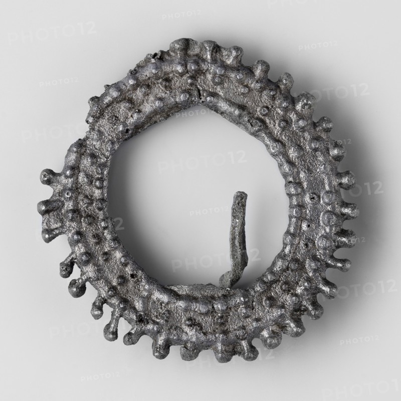Miniature brooch, votive offerings, wreaths, 1st–2nd century, Roman Empire 27 BC–476, toy, amulet; height 4.6 cm, width 0.9 cm, weight 0.018 kg, A roughly circular metallic object with a textured, bumpy surface and irregular protrusions around its outer edge, shown against a smooth light gray background; the object appears dark gray with silvery highlights and a slightly rough, pitted finish, and there is a thin, vertical metallic fragment rising from the inner edge.