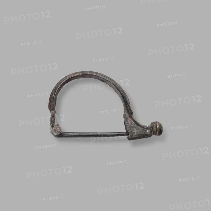 Aucissa-type fibula, Rome Empire, complete brooch with a flat ring decorated with an elongated roller. Earth-brown patina. Half of the thin roller, with a hole for attaching a needle, is broken off. End of the 1st century BC–1st century, Black Sea region. Fibula; height 3.2 cm, width 5 cm, depth 1.4 cm, weight 6 g., A small metal object shaped like an arch with a straight bar across its base sits centered against a plain light gray background; the metal shows a worn, textured surface with dark gray, brown, and greenish patina, and one end of the arch terminates in a rounded, slightly bulbous knob while the opposite end connects to a small rounded clasp.
