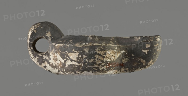 Oil lamp, unknown Gaulish workshop role/classification of maker or workshop uncertain, from 2nd century Gaul. Lamp from a mold. Round vessel with arms sloping outward, separated from the disc by a convex edge. Flat, concave disc with an almost centrally located oil hole. Two bars placed symmetrically on the arms; third vice placed vertically opposite the burner. Long wick channel decorated in the center with a wide flat groove bounded by two convex edges. Rounded end of the burner; large wick hole; no charring. Traces of a faded trademark on the bottom. Type: Buchi type Xa Loeschcke type X oil lamp. Dimensions: height 3.3 cm, width 11.8 cm, depth 6.6 cm., An object resembling a shallow bowl with a rounded handle and a circular hole is shown against a plain background; the surface is mottled with dark gray and black areas and worn patches of off-white and beige, with irregular flaking and rough texture visible across the form.