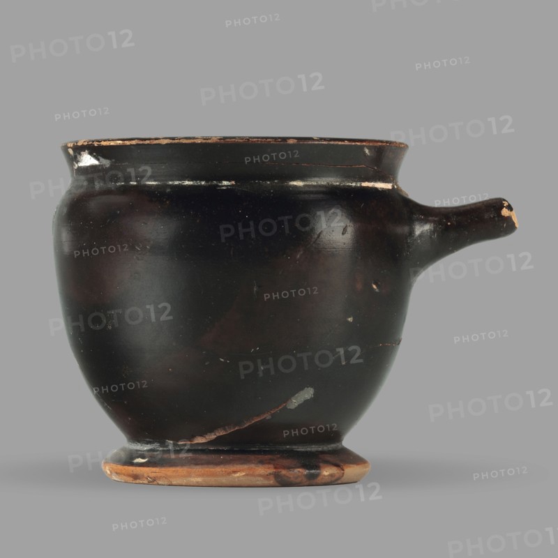 Askos with spout and strainer, unknown Attic workshop; Greek pottery toilet vessel with tubular spout and ribbon handle; top covered with a strainer; 5th century BC, Attica; vessel askos; height 6.2 cm, width 8.3 cm, depth 9 cm, weight 0.09 kg., A rounded ceramic vessel with a short spout on one side sits against a plain background; the vessel is primarily black with areas of dark brown and small patches where the surface shows lighter brown or beige; the rim and base display worn lighter brown edges, and there are a few small chips and scuffs revealing pale material beneath the dark surface; the shape includes a slightly flared rim, a bulbous body, and a low pedestal foot with mixed brown and beige tones.