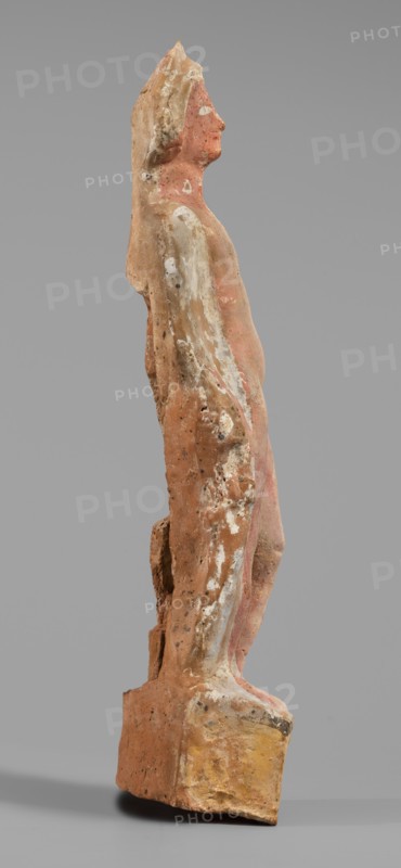 Male figurine, unknown Boeotian workshop. Standing man on a high quadrangular base, arched at the back. The weight of the body rests on the right leg, the left leg slightly forward. Torso naked at the front, leaning backward with the right hip protruding; slender body. A robe draped over the shoulders and covering the back conceals the hands. Head slightly tilted to the right; face treated schematically with large eyes. In the lowered right hand the figure holds an elongated object, while the left hand presses a round vessel to the chest. Base bears a visible strip of brown paint; the figurine shows traces of red-pink polychrome and the eyes were covered with white paint. 5th–4th century BC earlier dating: mid-5th century BC, Boeotia. Terracotta figurine; height 27 cm, width 6.8 cm, depth 6 cm, weight 0.31 kg., A weathered standing figurine viewed in profile on a rectangular base, rendered with an elongated torso and head looking forward; the surface shows worn patches of pinkish-red, ...