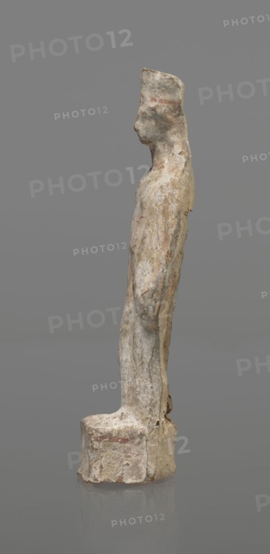 Female terracotta figurine of a standing woman, depicted frontally. Set on a small hollow, bottomless base, rectangular at the front and oval at the back. A large rectangular opening at the back shows clear traces of carving in the unfired clay. The woman stands with legs slightly apart, weight on the straight right leg, left leg bent and slightly forward. Arms lowered, slightly bent at the elbows, hands holding a cloak at hip level. Head turned slightly to the left on a long neck; elongated face with very faint features. Long combed-back hair flows over her shoulders. A polos on her head is decorated with a horizontal red line. Garments fit snugly with plastically defined folds. Traces of red polychrome on a white ground cover the entire surface; lines highlight garment folds and mark a necklace, and the belt tying the peplos. Damaged polychrome. 5th century BC, Greece. Height 19.6 cm., A weathered standing figure sculpture on a rounded rectangular base, shown in profile facing left, ...