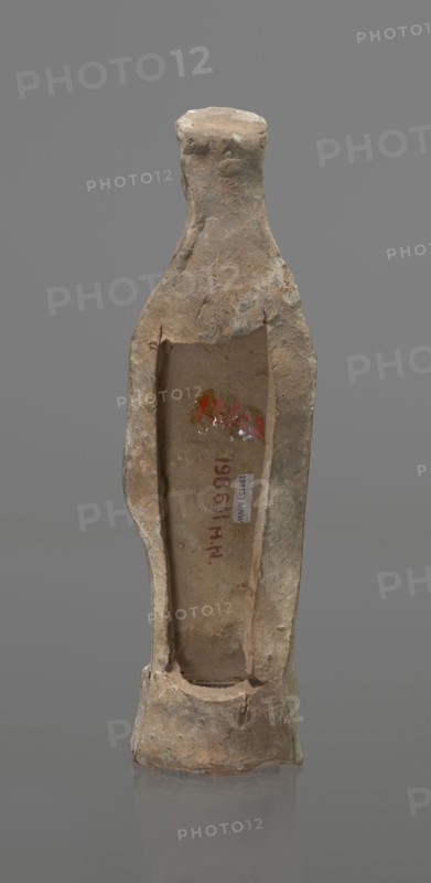 Female terracotta figurine of a standing woman, depicted frontally. Set on a small hollow bottomless base, rectangular at the front and oval at the back. A large rectangular opening at the back shows clear traces of carving in the unfired clay. The woman stands with legs slightly apart, weight on the straight right leg, left leg bent slightly forward. Arms lowered, slightly bent at the elbows, hands holding a cloak at hip level. Head turned slightly to the left on a long neck; elongated face with very faint features. Long combed-back hair flows over her shoulders. A polos on her head is decorated with a horizontal red line. Garments fit snugly with plastically defined folds. Traces of red polychrome on a white ground cover the entire surface; lines highlight the folds of the garment and mark a necklace, and the belt tying the peplos. Damaged polychrome. 5th century BC, Greece. Height 19.6 cm., A narrow, upright ceramic or clay vessel with rough, weathered surface and an open hollow fro...