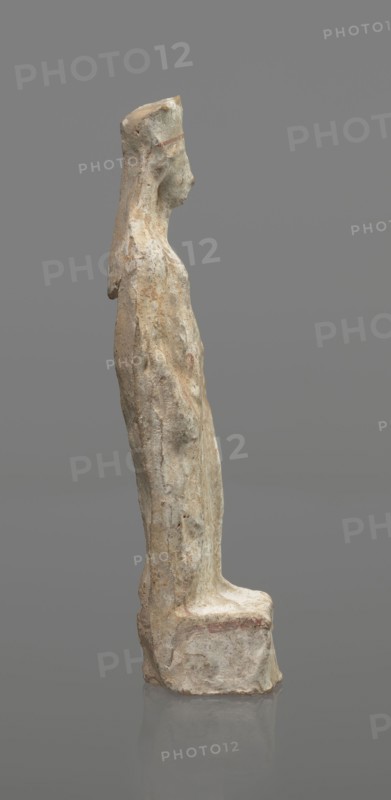 Female terracotta figurine of a standing woman, depicted frontally. Set on a small, hollow, bottomless base, rectangular at the front and oval at the back. A large, rectangular opening at the back shows clear traces of carving in the unfired clay. The woman stands with legs slightly apart, weight on the straight right leg; left leg bent and slightly forward. Arms lowered, slightly bent at the elbows, hands holding a cloak at hip level. Head turned slightly to the left on a long neck; elongated face with very faint features. Long, combed-back hair over the shoulders. Wears a polos decorated with a horizontal red line. Garments fit snugly with plastically defined folds. Traces of red polychrome on a white ground cover the entire surface; lines highlight the garment folds and mark a necklace, and the belt tying the peplos. Damaged polychrome. 5th century BC, Greece. Height 19.6 cm., A standing carved figure viewed in profile on a plain gray background, rendered in weathered pale stone ton...