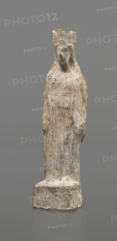 Female terracotta figurine of a standing woman, depicted frontally. Set on a small, hollow, bottomless base, rectangular at the front and oval at the back. A large, rectangular opening at the back shows clear traces of carving in the unfired clay. The woman stands with legs slightly apart, weight on the straight right leg, left leg bent slightly forward. Arms lowered, slightly bent at the elbows; hands hold a cloak at hip level. Head turned slightly to the left on a long neck; elongated face with very faint features; long combed-back hair over the shoulders. A polos on her head is decorated with a horizontal red line. Garments fit snugly with plastically defined folds. Traces of red polychrome on a white ground cover the entire surface; lines highlight garment folds and mark a necklace, and the belt tying the peplos. Damaged polychrome. 5th century BC, Greece. Height 19.6 cm., A standing sculpted figure on a rectangular base, rendered in weathered off-white and beige tones with areas o...