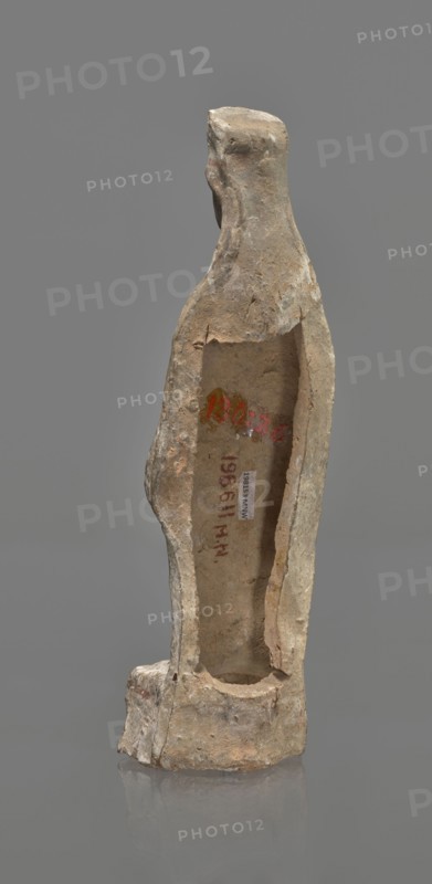 Female terracotta figurine of a standing woman, depicted frontally. Set on a small, hollow, bottomless base, rectangular at the front and oval at the back. A large rectangular opening at the back shows clear traces of carving in the unfired clay. The woman stands with legs slightly apart, weight on the right leg, left leg bent and slightly forward. Arms lowered, slightly bent at the elbows, hands holding a cloak at hip level. Head turned slightly left on a long neck; elongated face with very faint features. Long, combed-back hair flows over the shoulders. A polos on her head is decorated with a horizontal red line. Garments fit snugly with plastically defined folds. Traces of red polychrome on a white ground cover the entire surface; lines highlight garment folds and mark a necklace, and the belt tying the peplos. Damaged polychrome. 5th century BC, Greece. Height 19.6 cm., A weathered, narrow vertical sculpture with an elongated neck and simplified head stands against a neutral gray b...