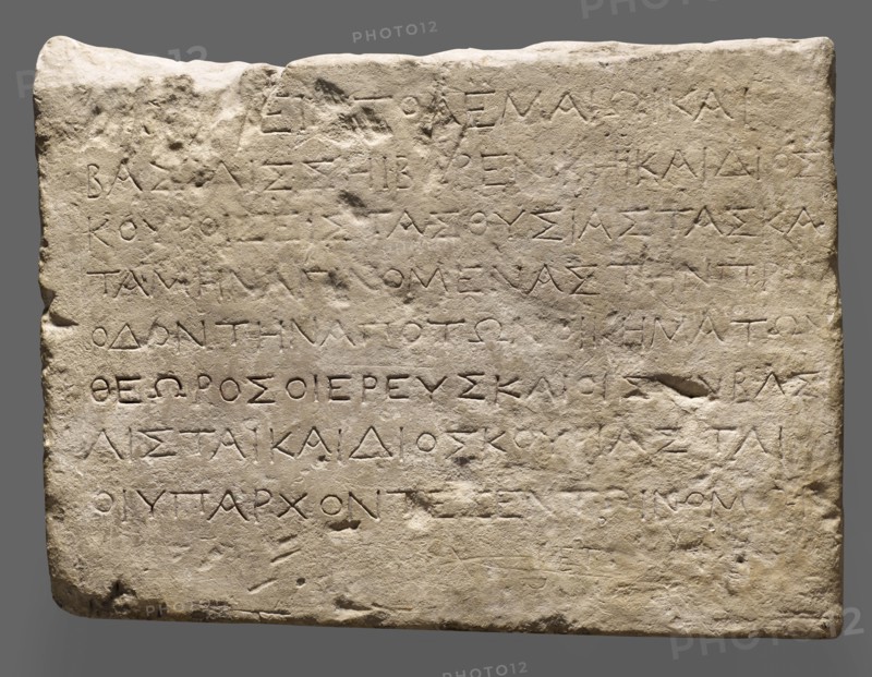 Plate with a Greek dedication to Ptolemy III, Berenice and the Dioscuri;, Greek inscriptions; Ptolemy III Euergetes ruler of Egypt, 246–221 BC; dedications; dated 246–241 BC before 241 BC, no epithet theoi Euergetai; Egypt; height 30.2 cm, width 40.5 cm, depth 7.7 cm, weight 15.728 kg, A rectangular stone slab with an uneven, worn surface and rounded corners rests against a neutral gray background; the slab is light beige with areas of darker beige and pale gray discoloration, and it shows surface pitting, chips, and shallow grooves across its face. The top edge is irregular and slightly raised in places, and there are a few shallow depressions and rough patches scattered across the surface. The overall texture appears weathered and slightly grainy, with subtle shadows emphasizing the carved indentations and surface irregularities.