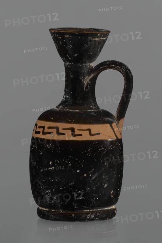 Black-finned aryballois lekythos, /workshop., repossession Silesia. Lekythos, black-finned pottery. Large, ring-shaped foot. Body squat, barrel-shaped with rounded shoulders. Neck narrow, clearly separated from body, with flaring rim. Upper part of body has a slightly irregular clay-colored orange band not covered with finnis; on it, at the front, a rather carelessly executed meander interrupted with black paint. Upper part of the vice is tilted to the left. Condition: minor chips, rim edge worn. Date: 1st half of the 19th century; 4th century BC. Unknown site/place of origin. Vessel lekythos. Height 10.9 cm; diameter 5.4 cm., A dark ceramic vessel with a narrow flared neck and a rounded body, featuring a single curved handle attaching from the upper neck to the shoulder; the surface shows worn spots and flecks revealing lighter underlying material, and a horizontal band of lighter color encircles the shoulder with a dark zigzag motif on it; visible colors include black, a tan or light...