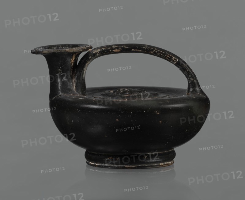 Black-finished askos, unknown Campanian workshop, black-finished pottery, askos, Campania Italy. Askos with a profiled foot and a semicircular body closed at the top with a slightly recessed surface. Narrow neck ending in a wide rim. Ribbon-shaped handle attached to the neck and to the upper surface of the vessel. Appliqué with a female head glued to the upper surface of the vessel, probably Medusa, surrounded by a wreath of intertwined snakes. Finish is black, shiny, worn in places, particularly on the handle and the edge of the rim. Light orange clay. Handle and neck cracked and glued. Mid 3rd century BCE., A rounded ceramic vessel with a single arched handle and a short spout, shown against a plain gray background; the surface of the vessel is predominantly black with areas of worn paint revealing lighter, tan-colored spots and edges, and there are small scuffs and chips across its body and handle.